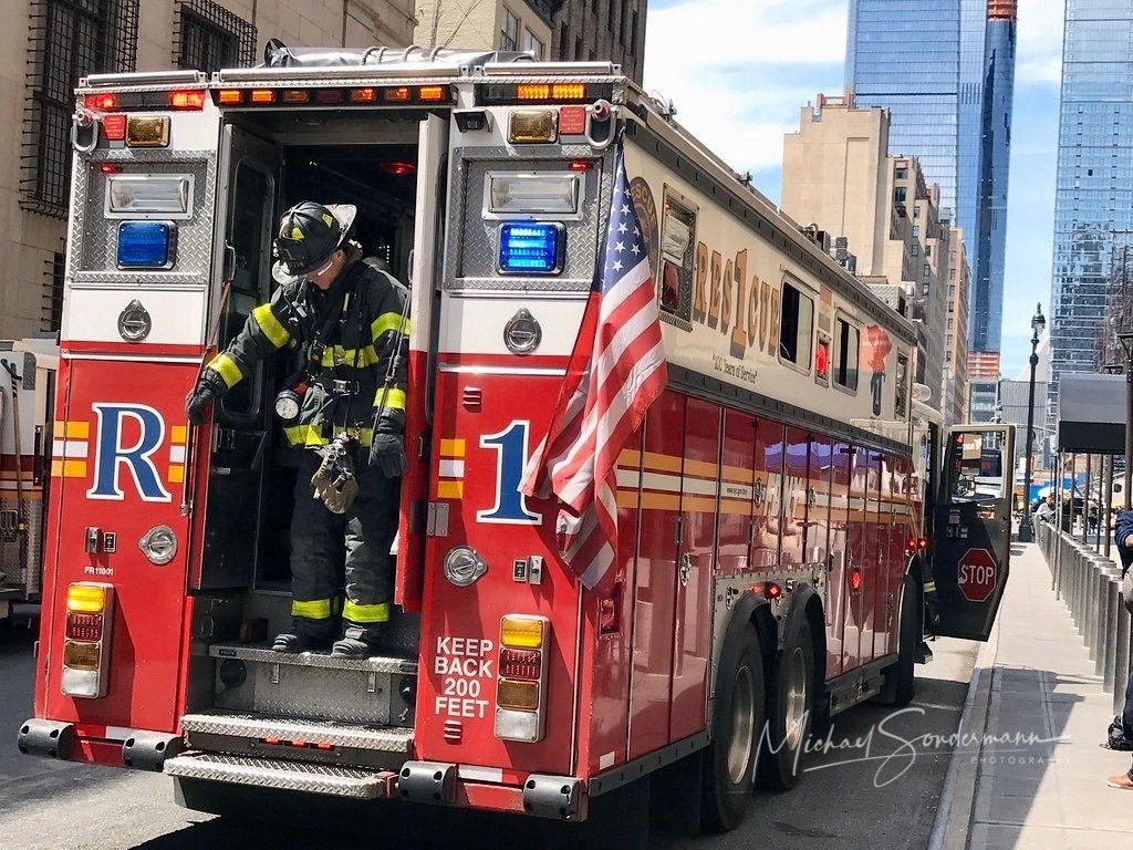 Ein roter Feuerwehrtruck der New Yorker Feuerwehr steht gerade mit Blaulicht bei einen Einsatz in die USA Fahne weht im Wind.