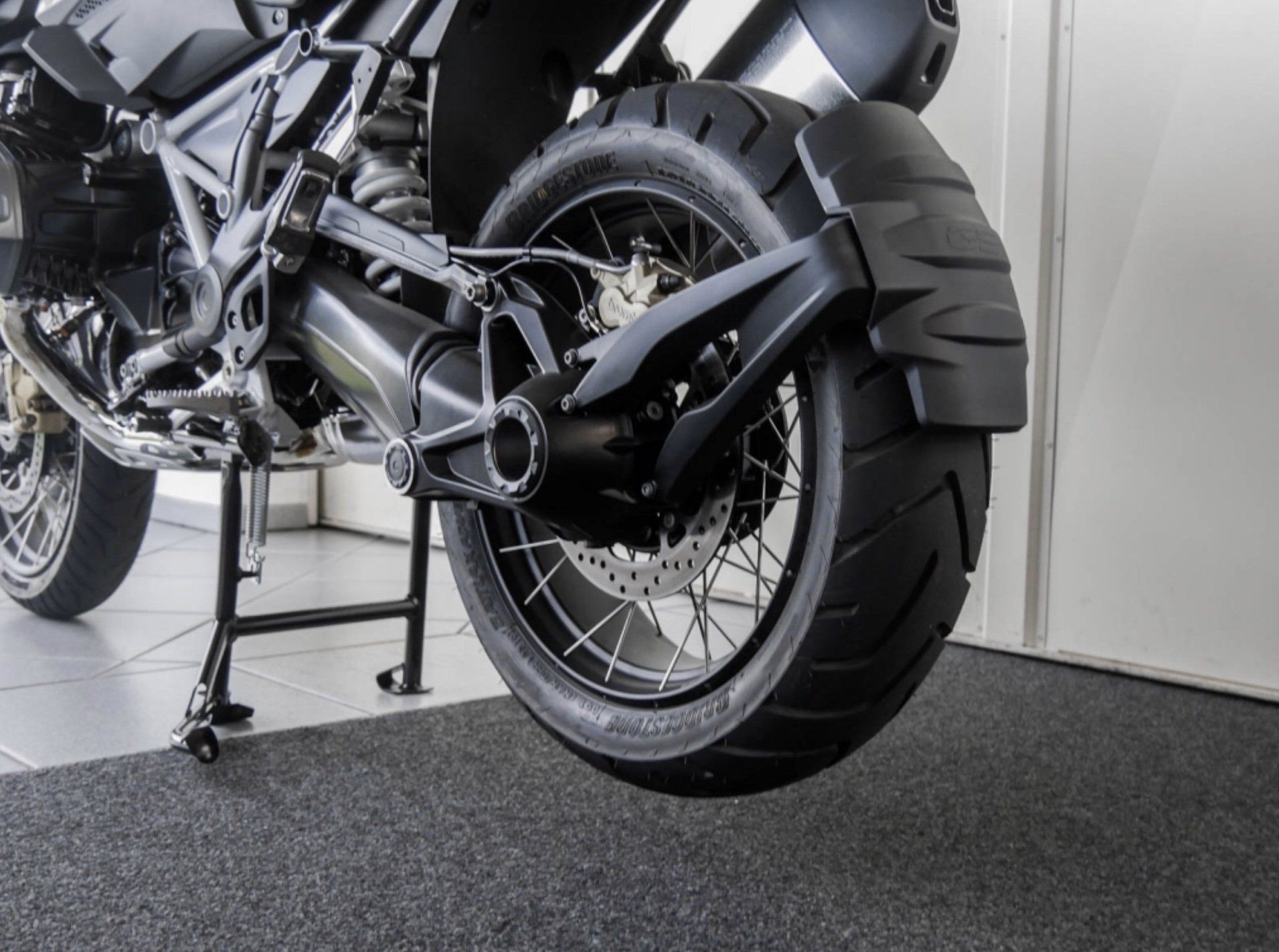 Ein Motorrad steht auf Bodenfliesen in der Sicht auf die hintere Felge vor einer weißen Wand auf dem Ständer.