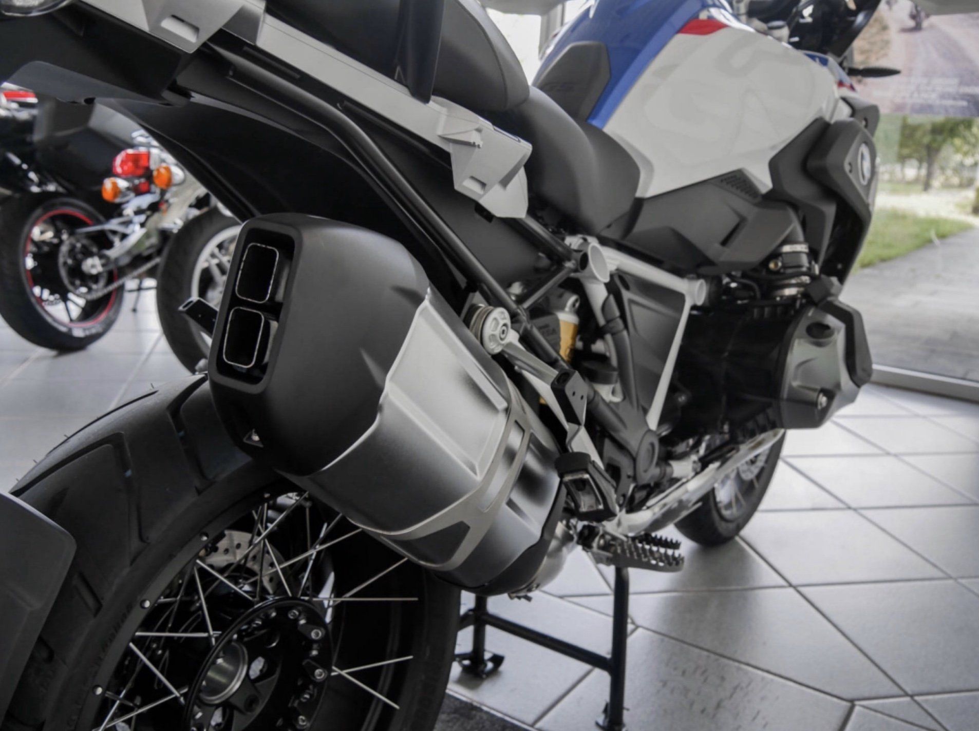 Ein Motorrad steht auf Bodenfliesen in der Sicht auf die Sportauspuffanlage vor einer weißen Wand auf dem Ständer