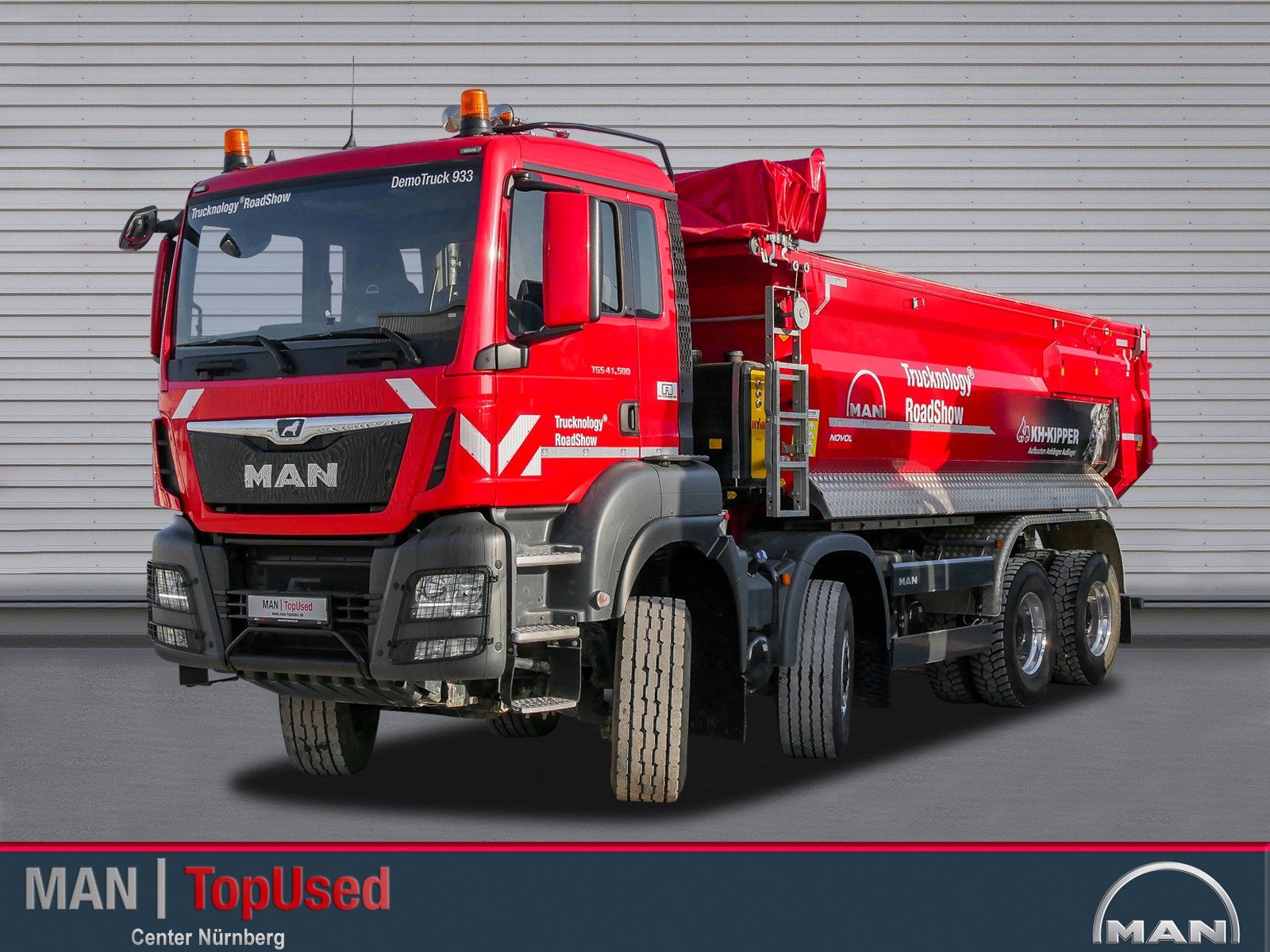 Ein großer roter 4 achsiger MAN Truck von vorne rechts gesehen mit eingeschlagenen Vorderrädern vor einer geriffelten Wand.