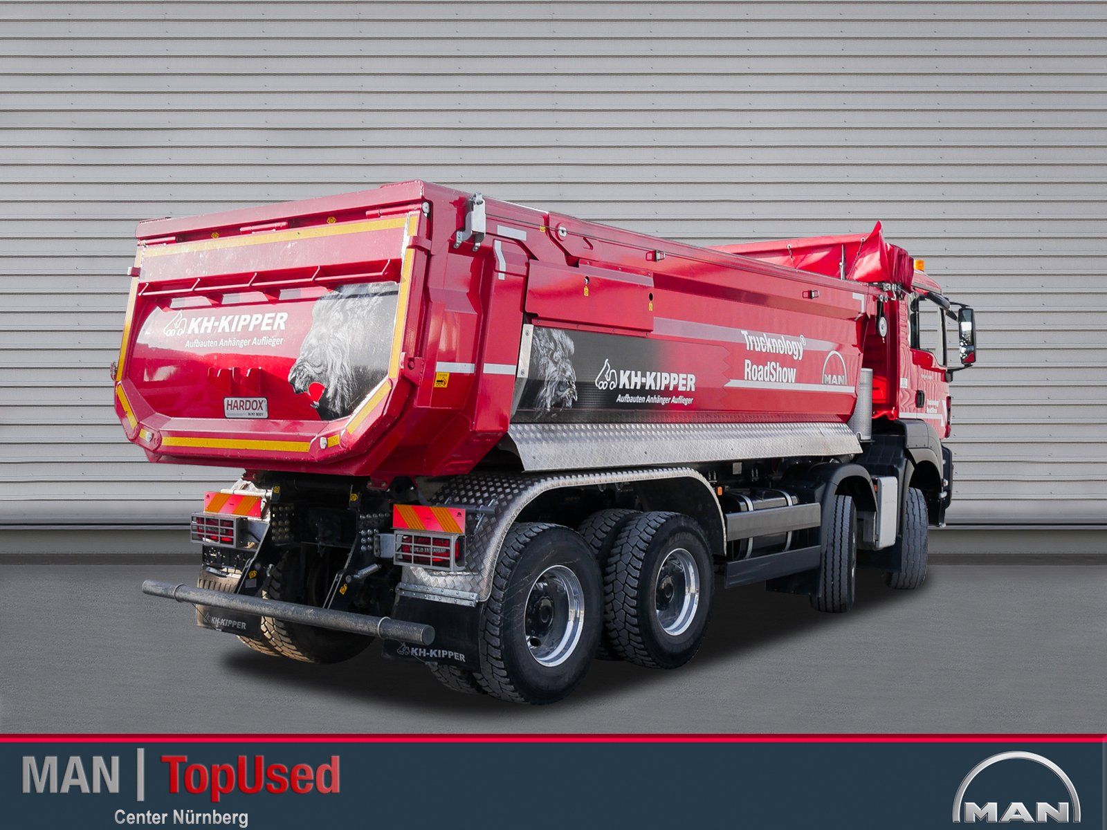 Ein großer roter 4 achsiger MAN Truck von hinten rechts gesehen mit eingeschlagenen Vorderrädern vor einer geriffelten Wand.