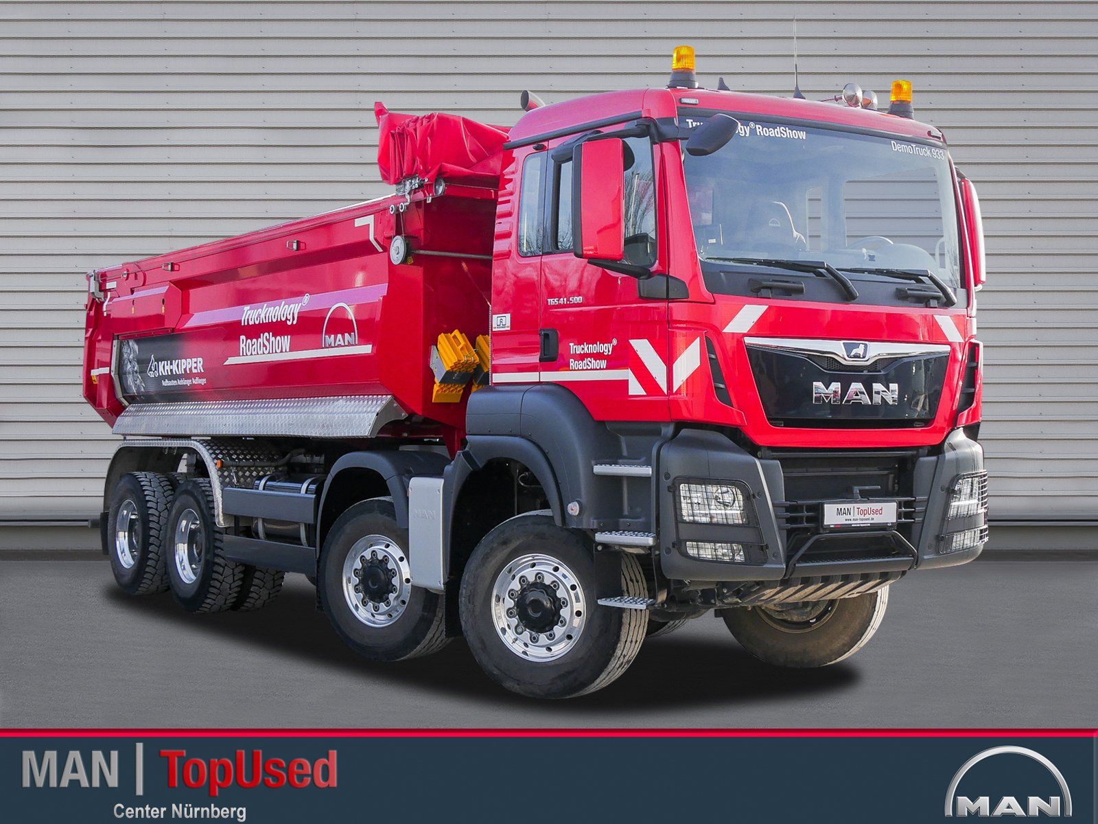 Ein großer roter 4 achsiger Truck von vorne links gesehen mit eingeschlagenen Vorderrädern vor einer geriffelten Wand.