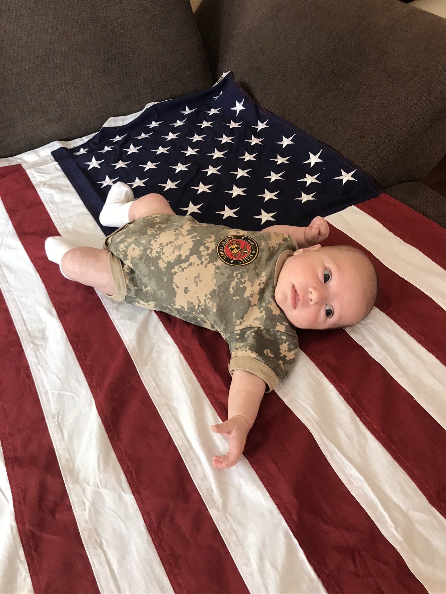 Ein Baby liegt in einem Strampler in Tarnfarben auf der USA Fahne auf dem Sofa und guckt in die Kamera.