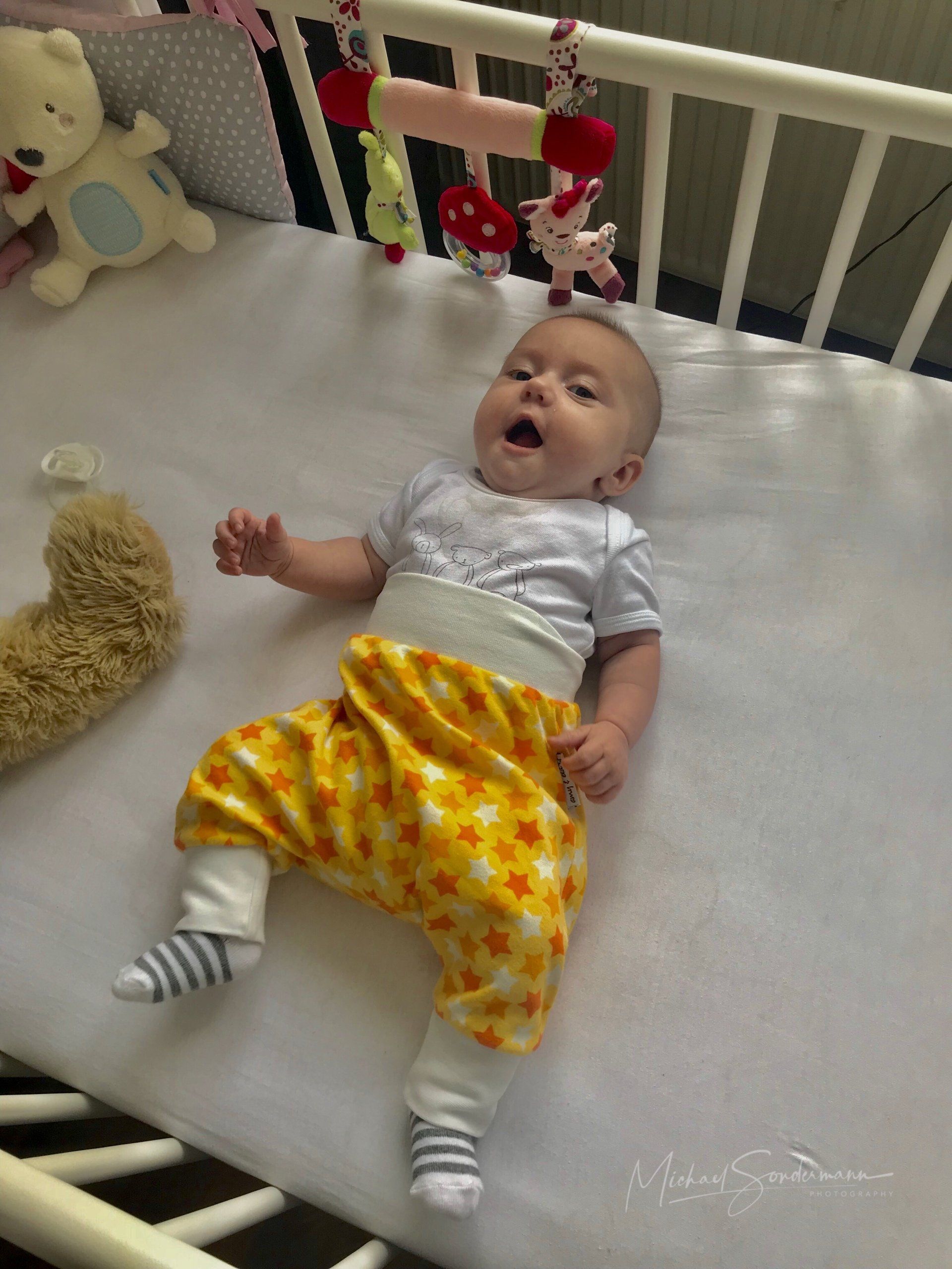 Ein Baby liegt mit einer gelben Hose gähnend im Kinderbett.