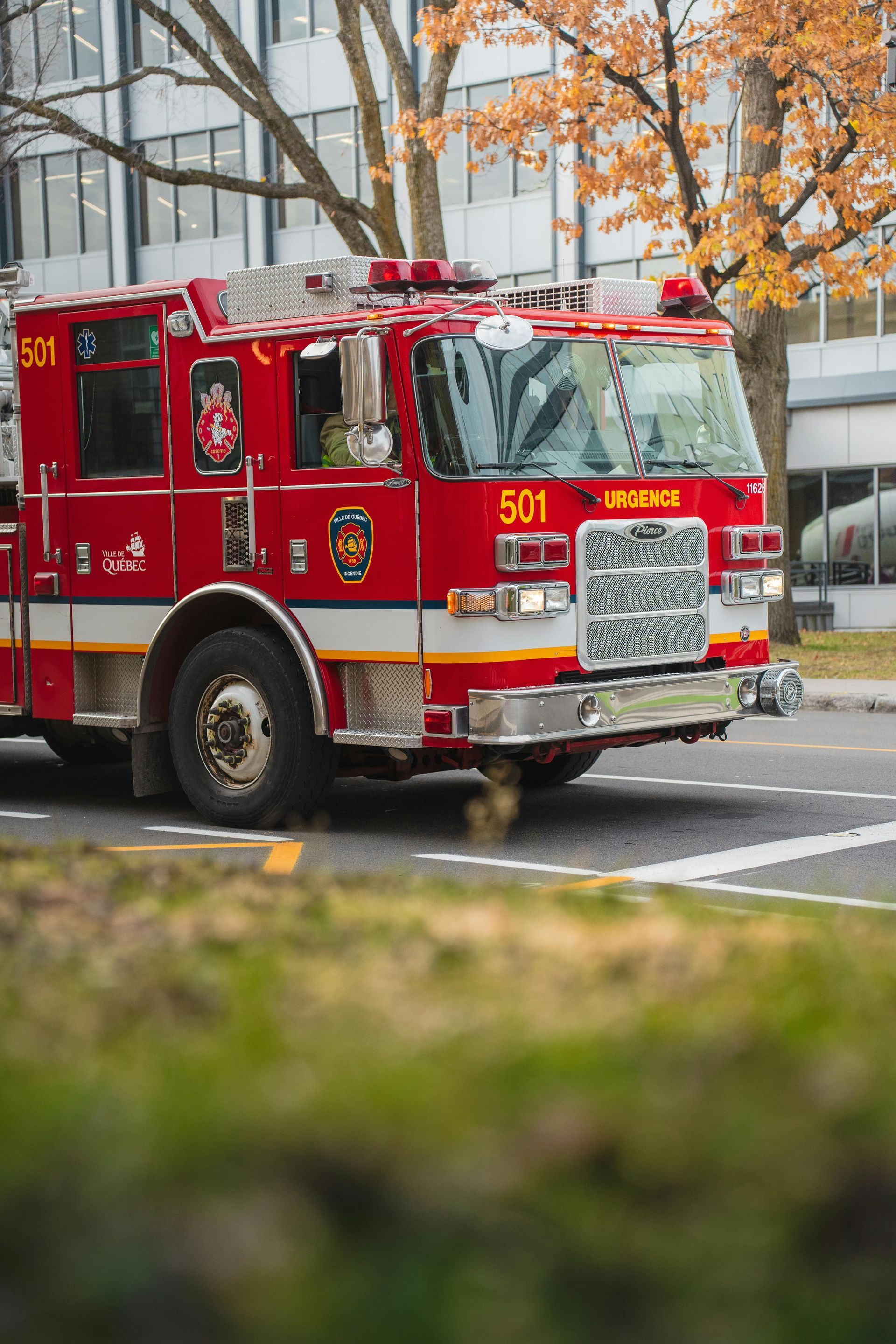 Red fire truck on a city street with a building and fall foliage in the background.