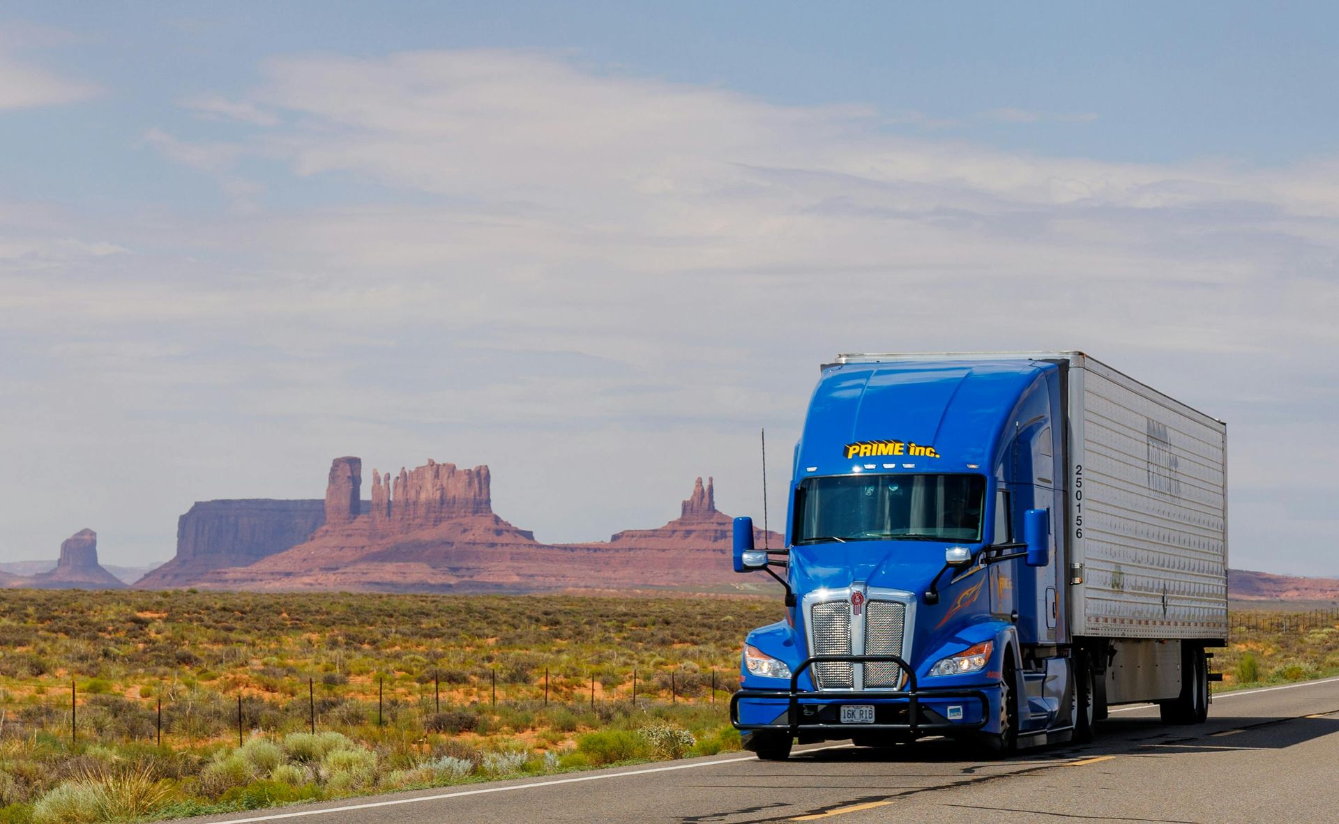Blue semi-truck driving on a desert highway with red rock formations in the background.