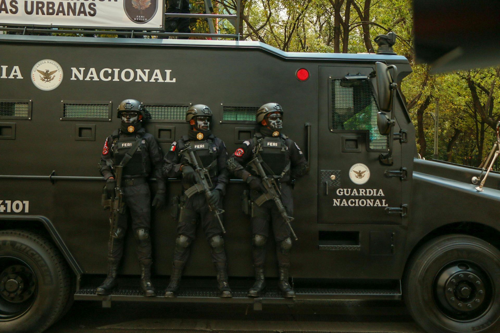 Three uniformed National Guard members stand in front of an armored vehicle, holding rifles.