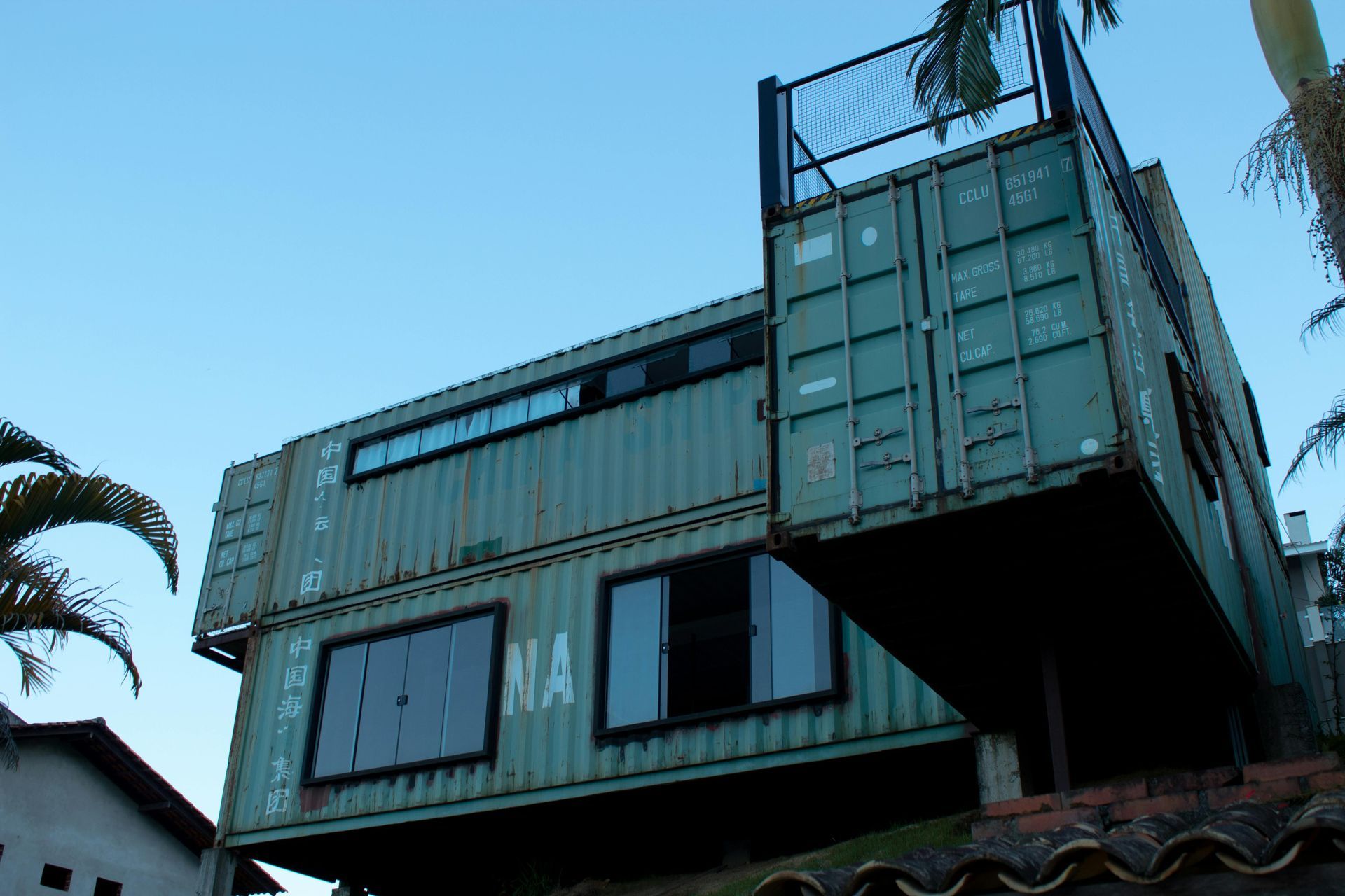 Two-story house made of green shipping containers with windows and a rooftop deck.