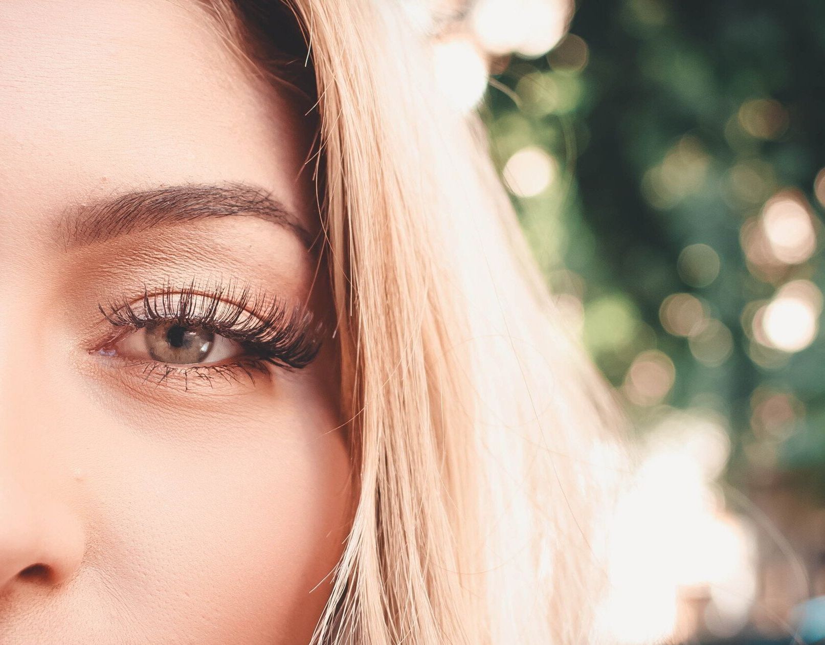Nahaufnahme des Gesichts einer Frau mit blondem Haar, markanten Wimpern und goldenem Lidschatten, die vor einem unscharfen grünen Hintergrund nach rechts blickt.