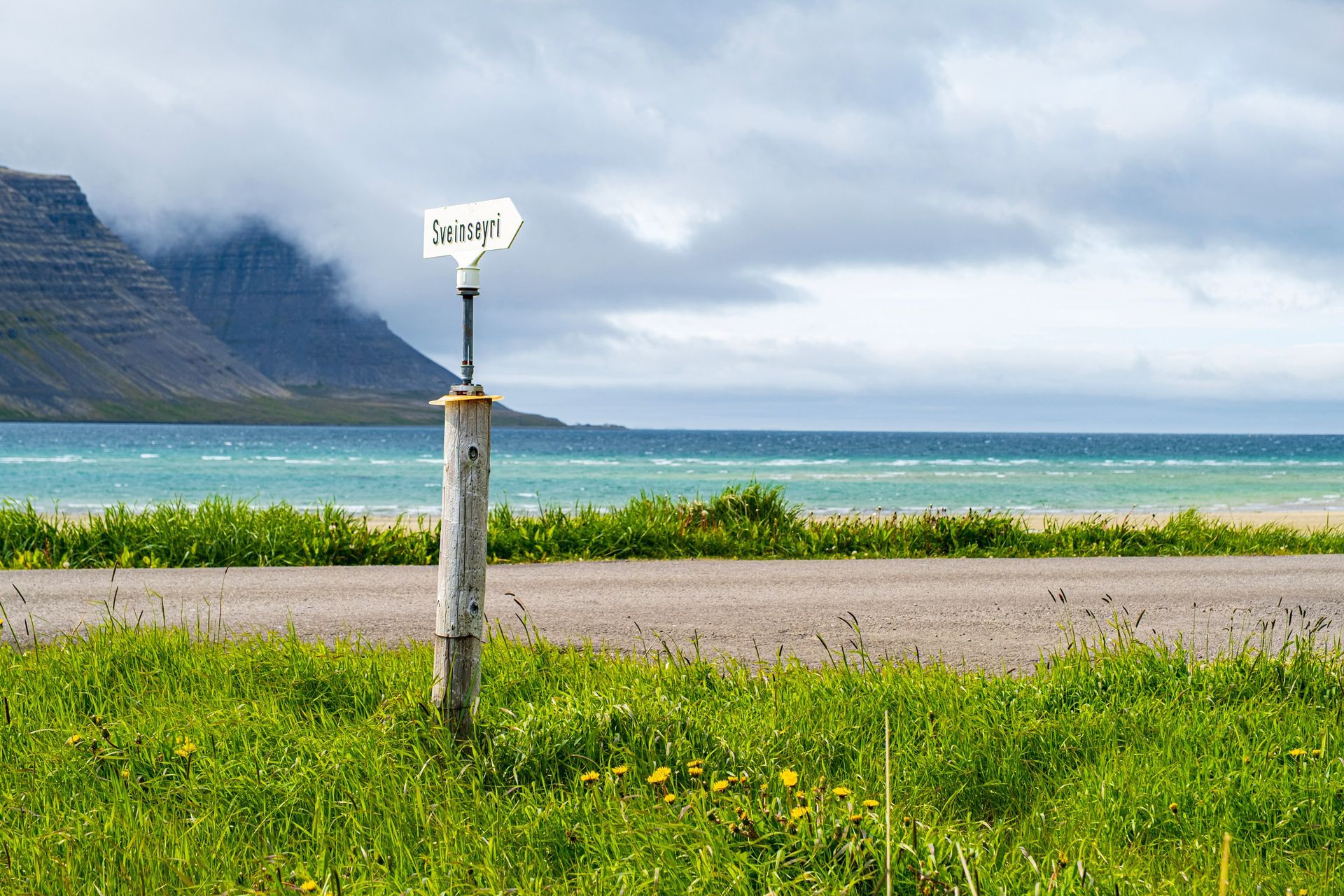 Roadside sign points toward turquoise water with mountains in background. Overcast sky.