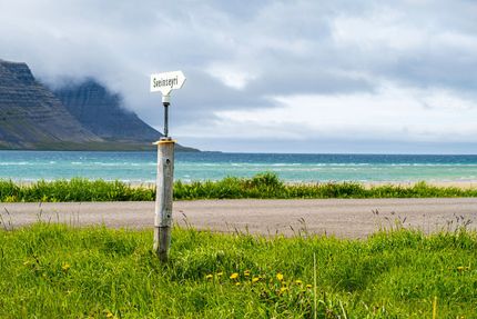 Roadside sign points toward turquoise water with mountains in background. Overcast sky.