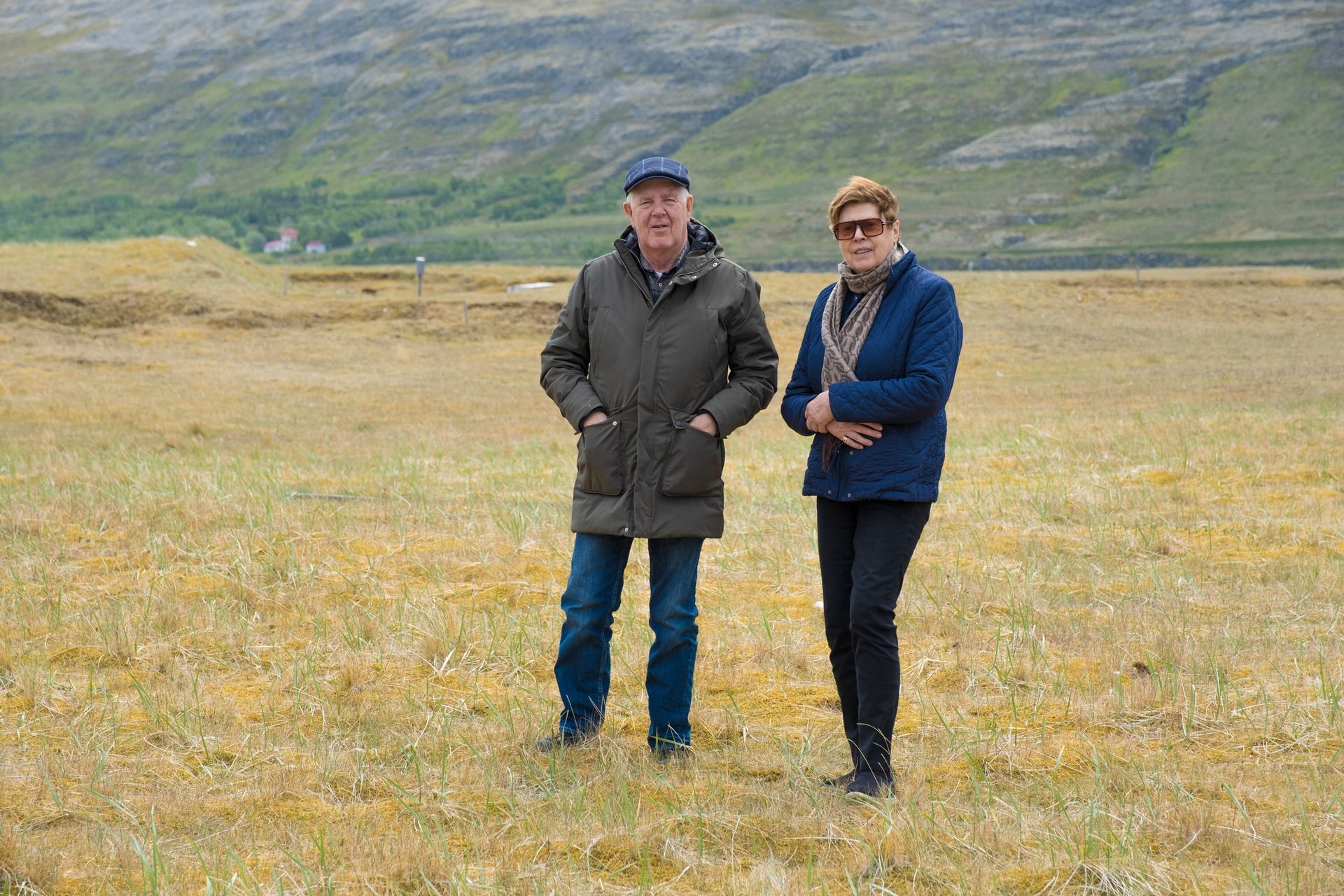 Two people standing in a field, mountains in the background. One in a dark coat, the other in a blue jacket.