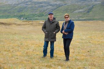 Two people standing in a field, mountains in the background. One in a dark coat, the other in a blue jacket.