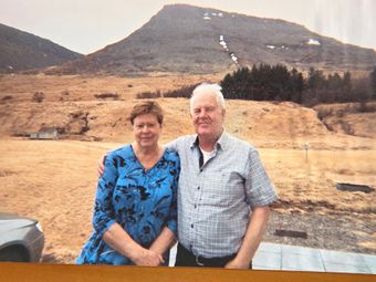 Couple poses outdoors in front of a mountain. Woman in blue floral top, man in a plaid shirt, both smiling.