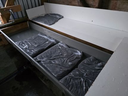 Bags of dark material in a water-filled trough, set inside a white-walled container.