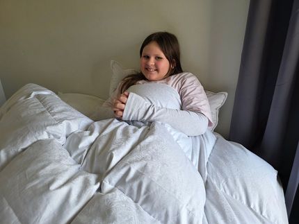 Girl smiles, nestled in bed under a fluffy white comforter, holding a pillow. Grey curtain background.