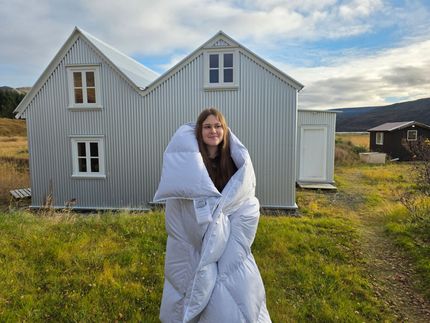 Person wrapped in white comforter stands in front of a metal building on a grassy field with a dark cabin in the background.