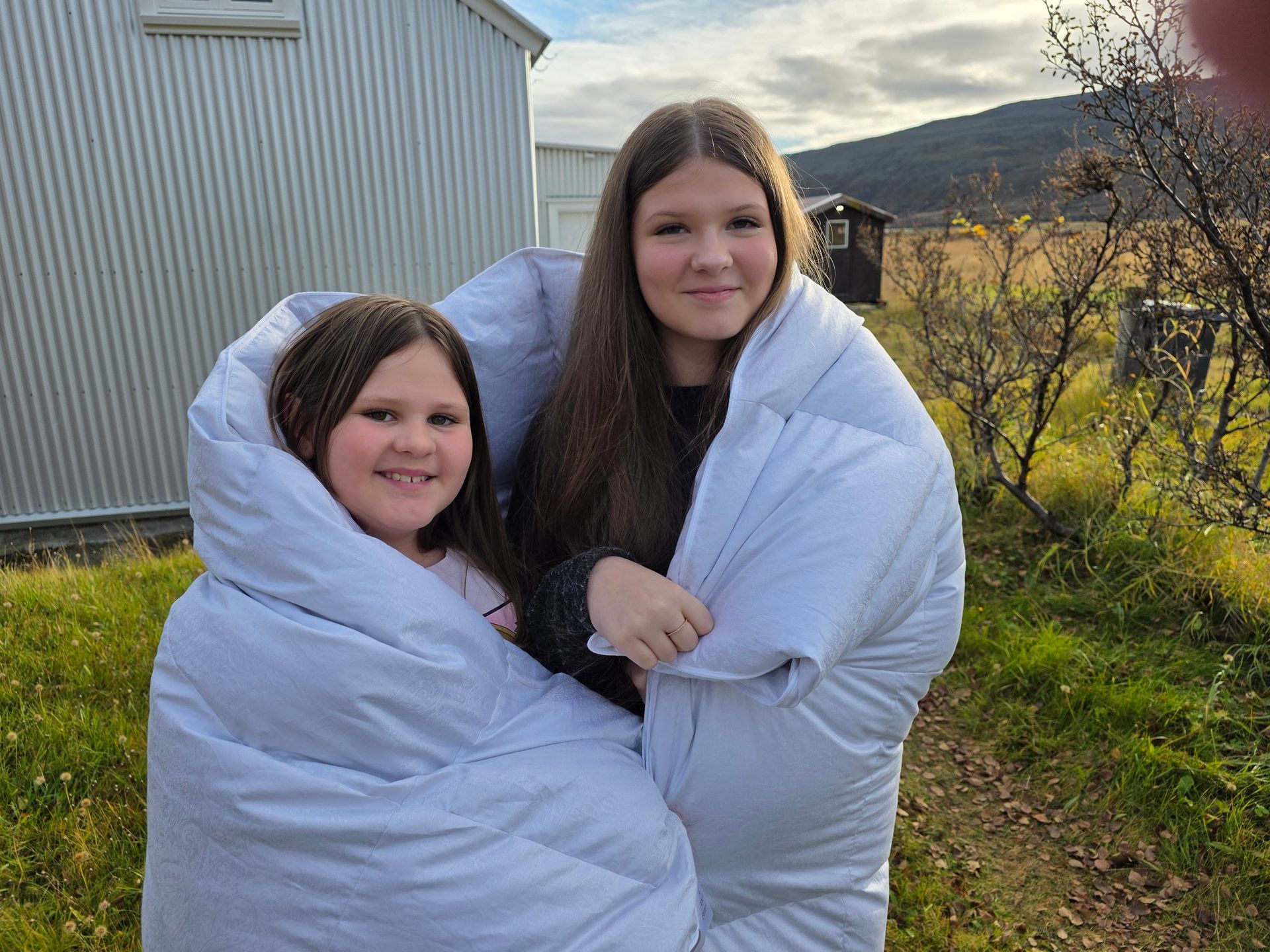 Two girls wrapped in a large, light blue blanket, smiling outside near a metal building.