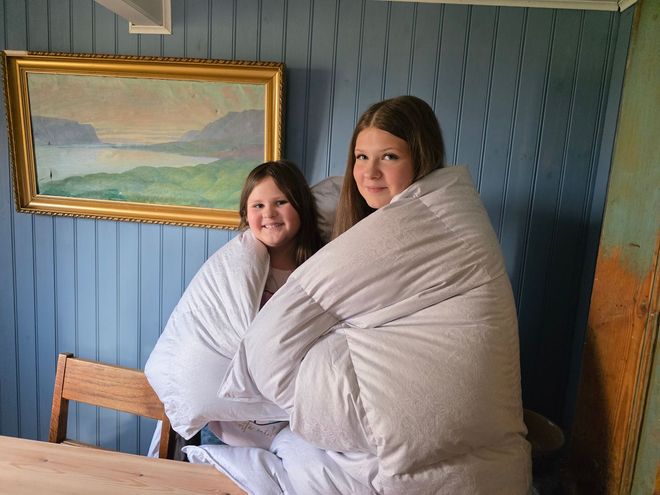 Two people wrapped in a white blanket, smiling, in front of a painting. Blue wall, wooden table.