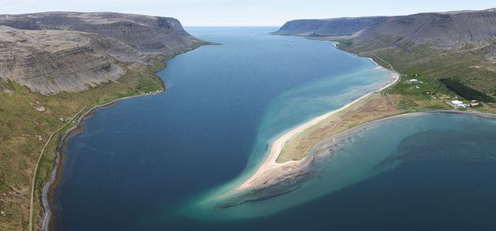Aerial view of a narrow, sandy peninsula dividing a wide blue lake, flanked by gray cliffs under a clear sky.