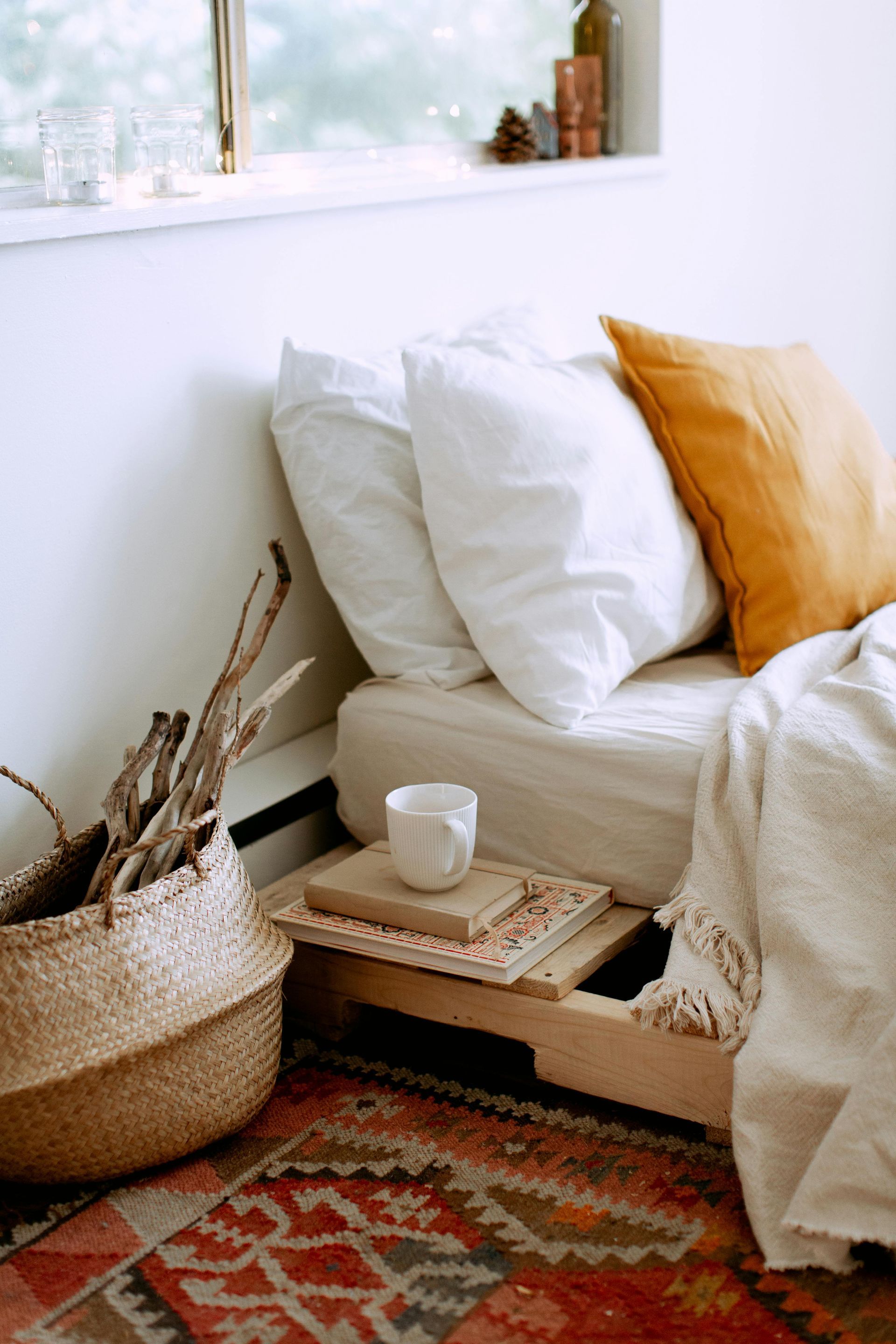 Cozy bedroom corner with bed, pillows, blanket, basket, and wooden shelf holding a cup and books.