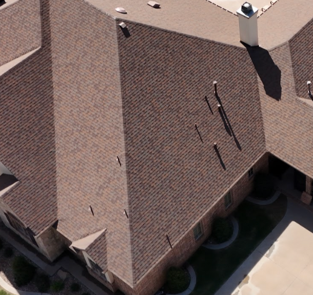 An aerial view of a brown-roofed home with a pool, a large concrete driveway, and a fenced backyard with raised gardens.