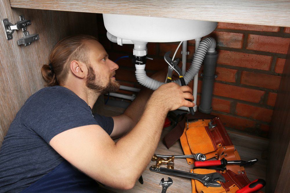 A Man is Fixing a Sink Under a Cabinet — Tony Brown Plumbing & Gas Services in South Grafton, NSW