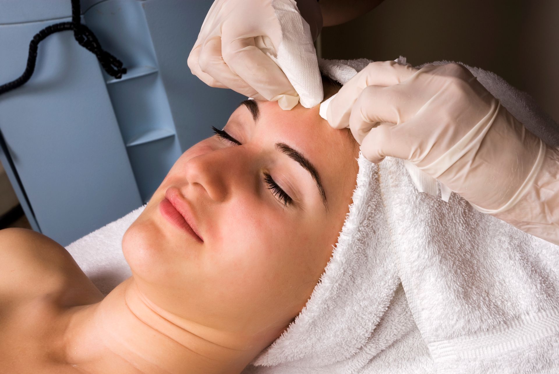 A person receiving a facial treatment, lying with eyes closed, hands in gloves on forehead.