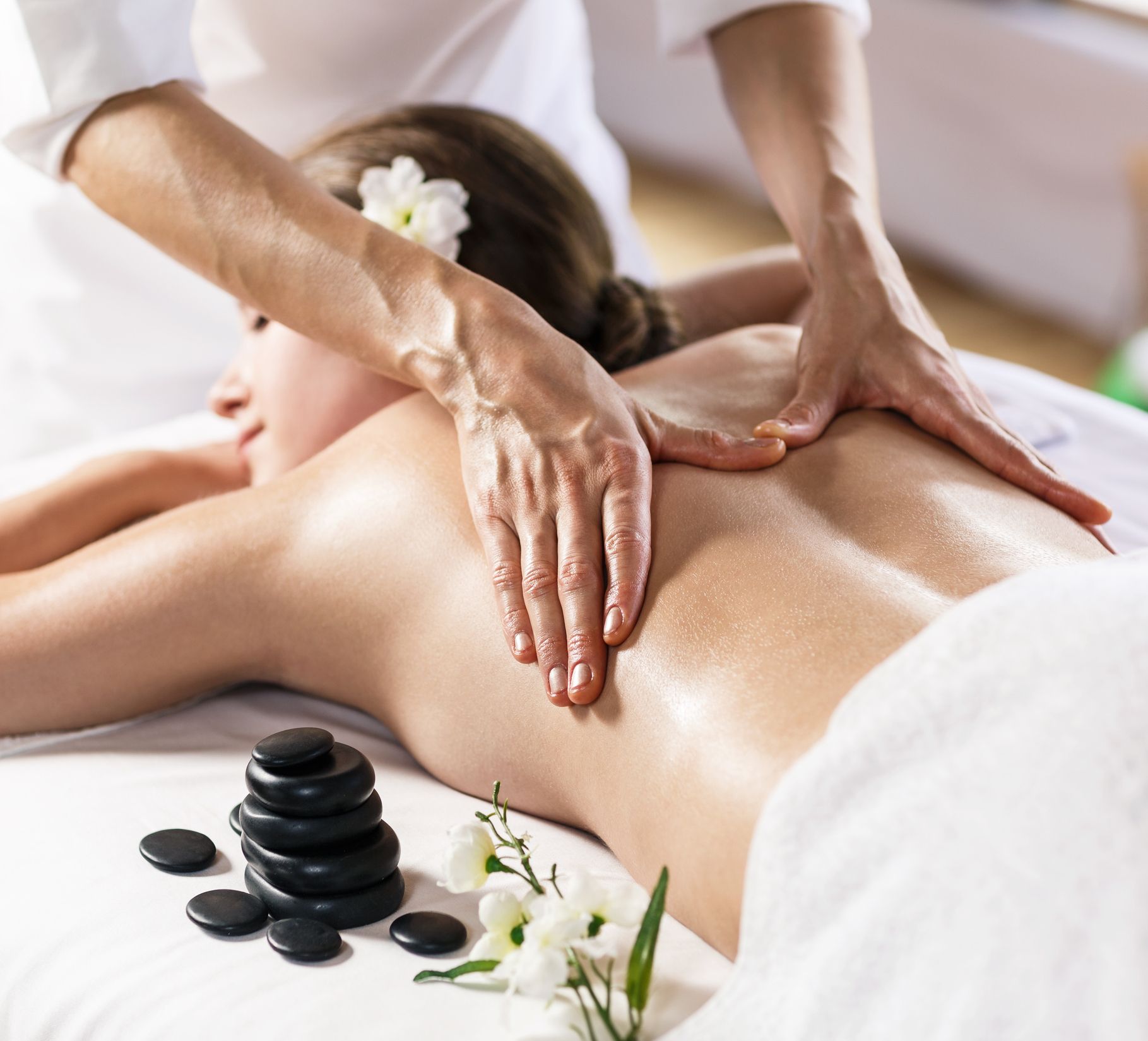 Person receiving back massage; therapist's hands on bare back, hot stones nearby, white flowers.