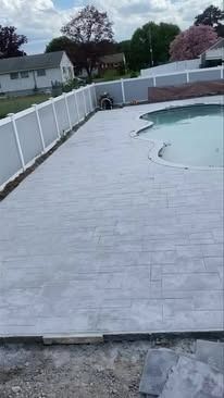 A concrete patio next to a swimming pool with a white fence.