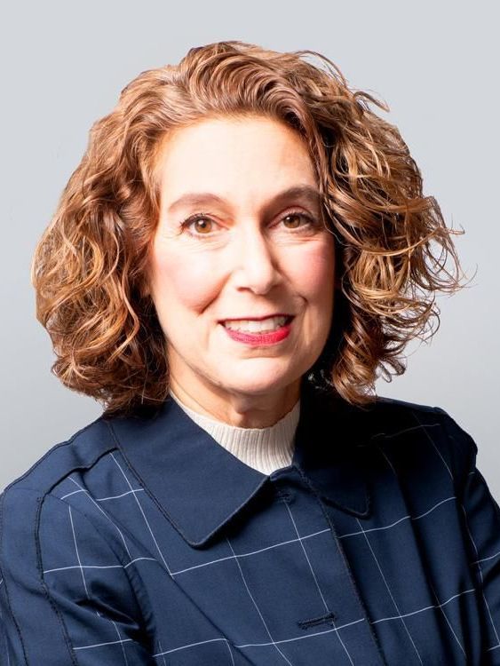 A woman with curly hair is wearing a blue jacket and smiling.