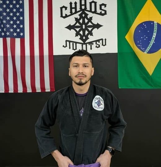 Man in black Jiu-Jitsu gi with purple belt, standing in front of flags.