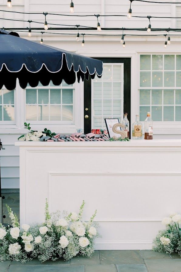 A white bar with a blue umbrella and flowers in front of a white house.