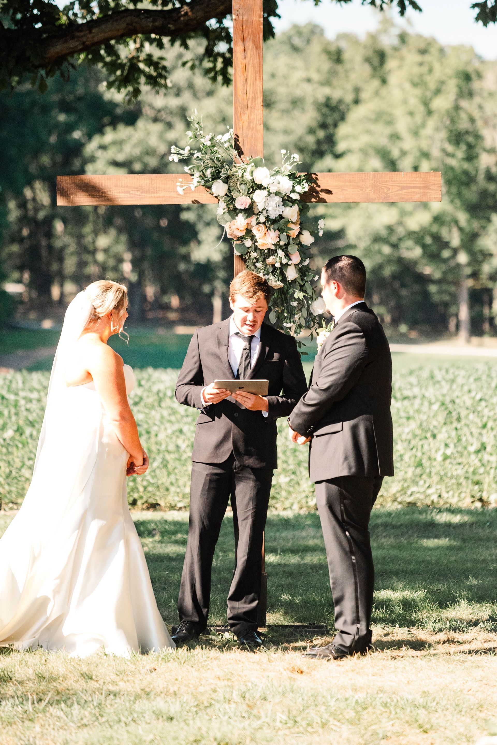 A bride and groom are standing in front of a wooden cross during their wedding ceremony.