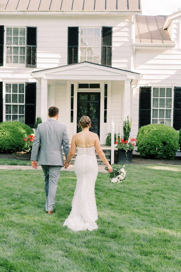 A bride and groom are walking in front of a white house holding hands.