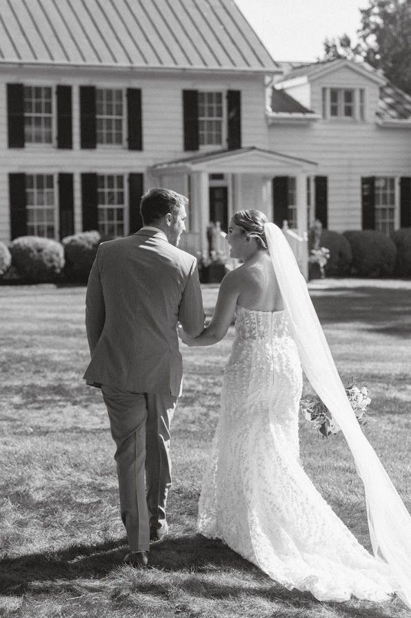 A bride and groom are walking in front of a large white house holding hands.