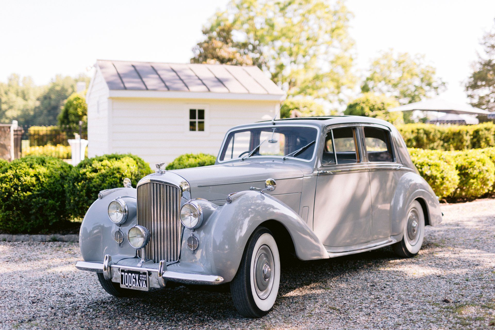 An old gray car is parked in a gravel driveway in front of a white house.
