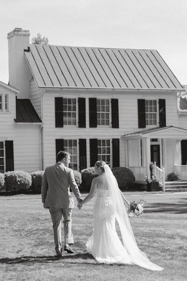 A bride and groom are walking in front of a large white house holding hands.