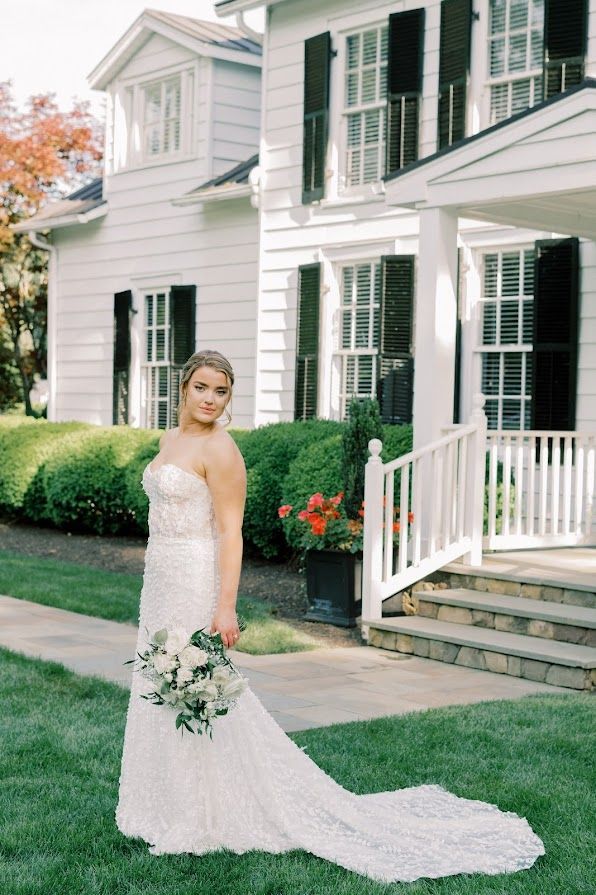 A bride in a wedding dress is standing in front of a white house holding a bouquet of flowers.
