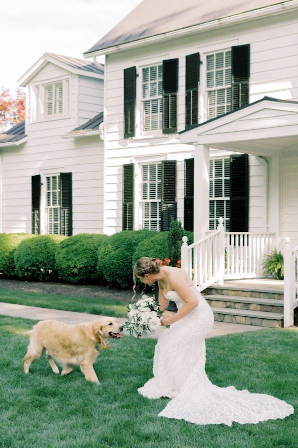 A bride in a wedding dress is playing with a dog in front of a white house.