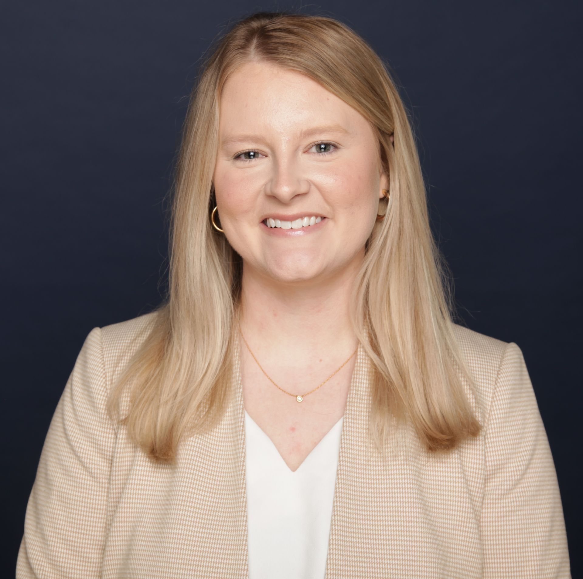 Woman with blonde hair wearing a beige blazer smiles in a studio setting.