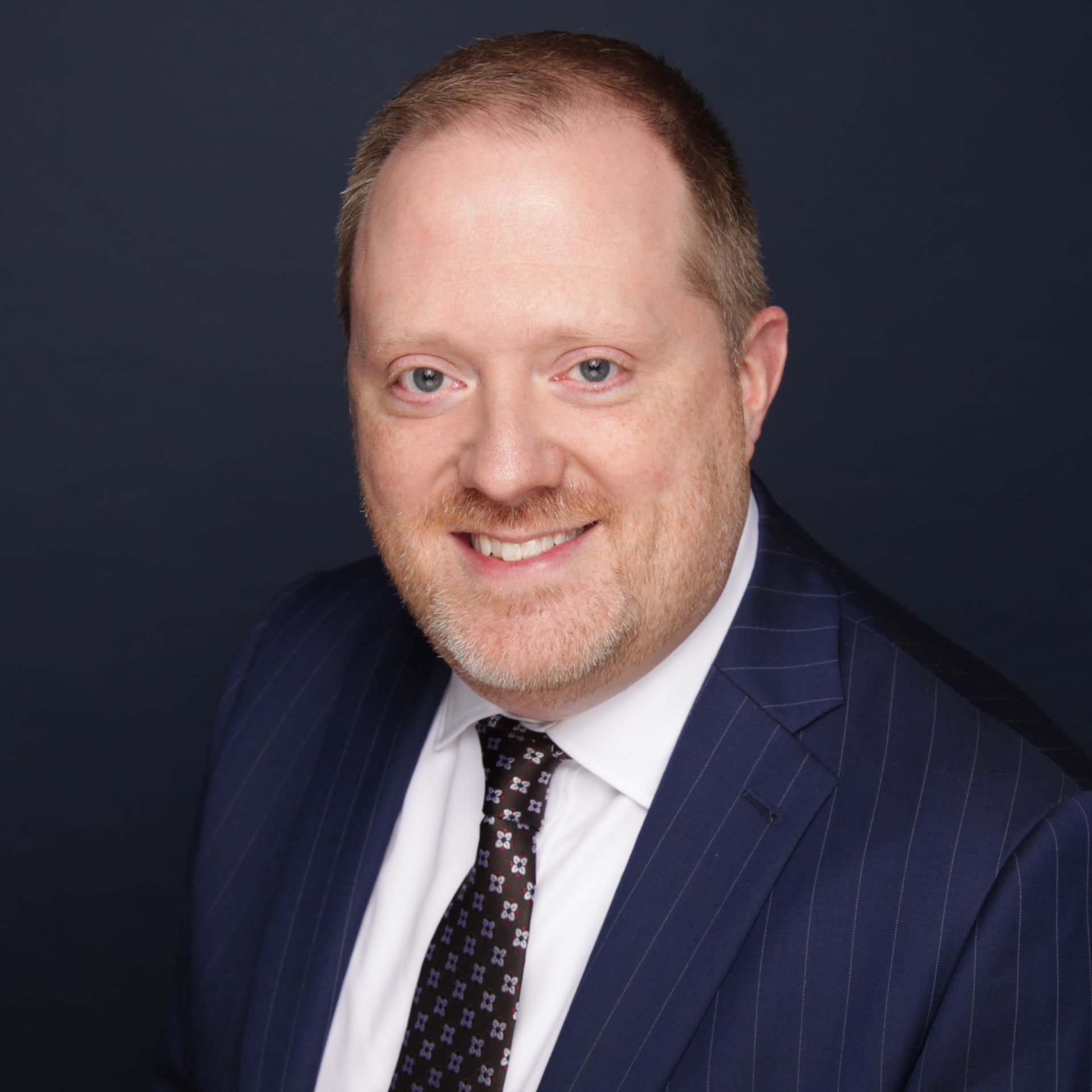 Man in pinstripe suit, smiling, with a patterned tie against a dark blue background.
