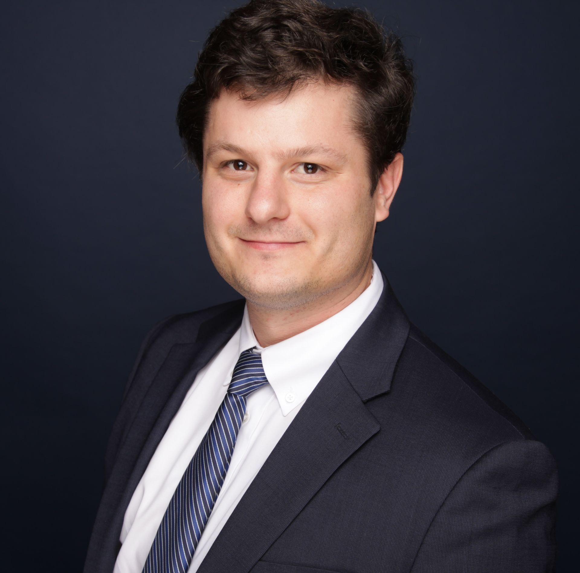 Man in a suit smiles, dark hair, blue tie, against a dark blue background.