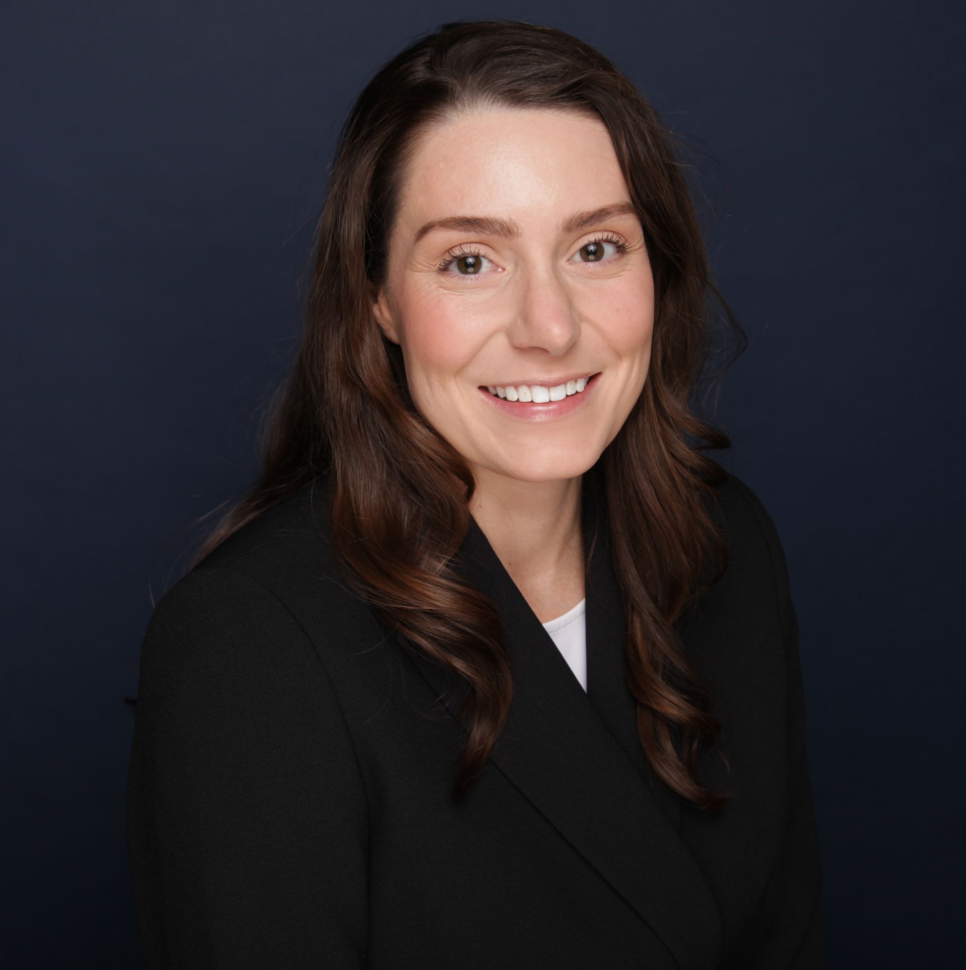 Woman with long brown hair smiling, wearing a black blazer and white shirt, against a dark blue background.
