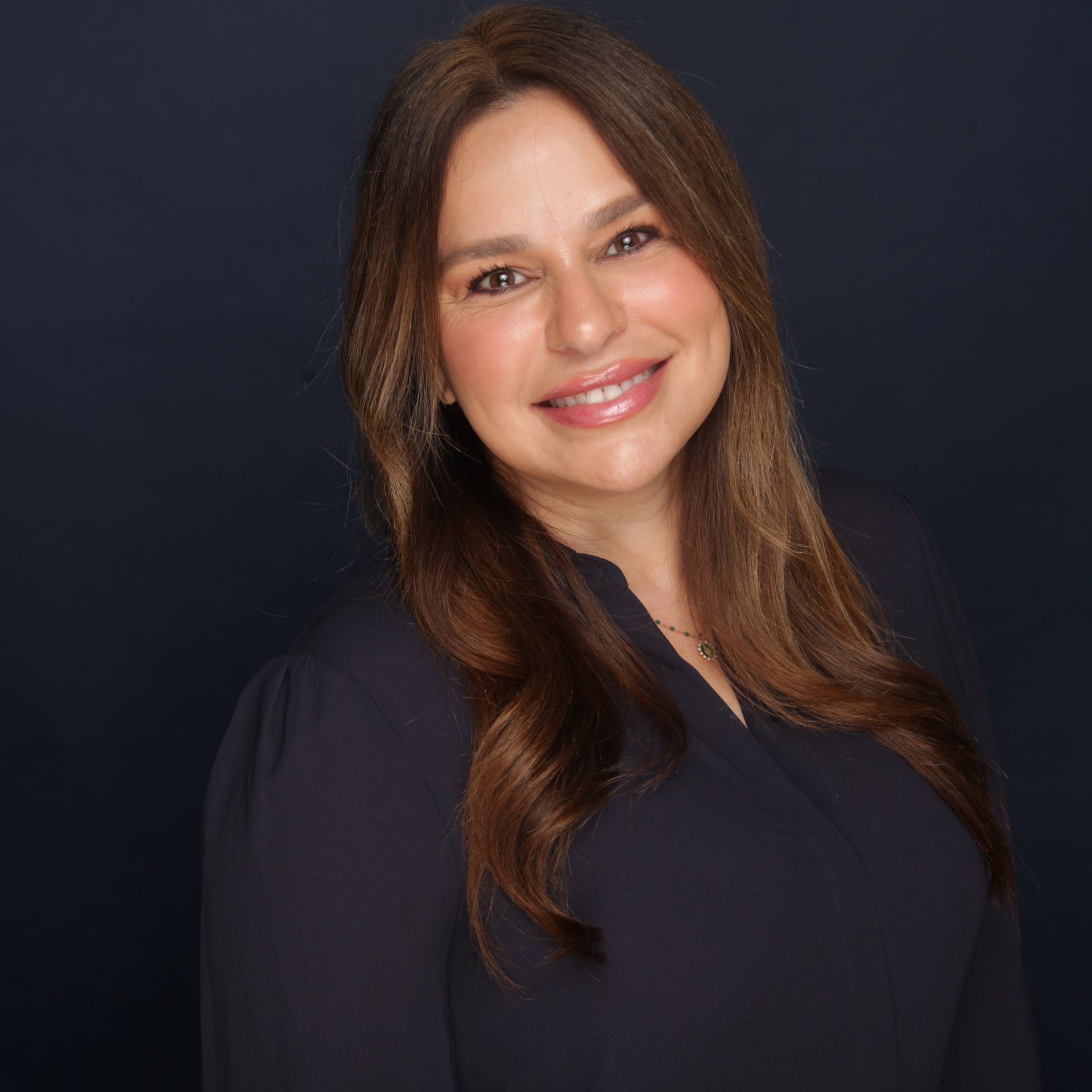 Woman with long brown hair, smiling, wearing a dark blue blouse against a dark background.