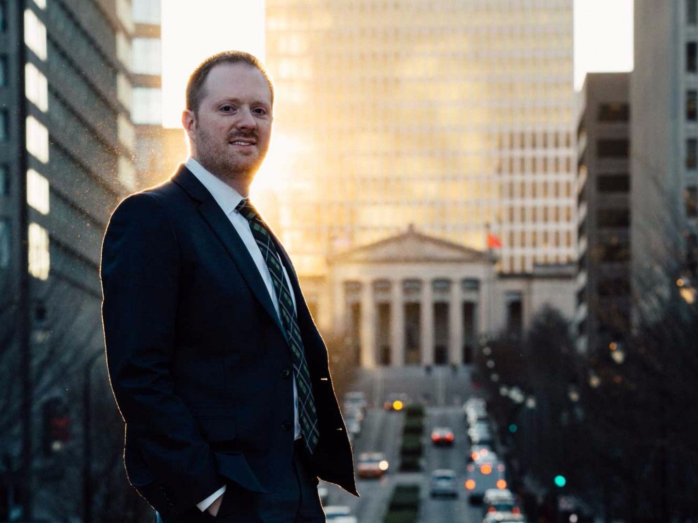 Man in suit stands on city street, looking at the camera. Tall buildings in the background with sunlight.