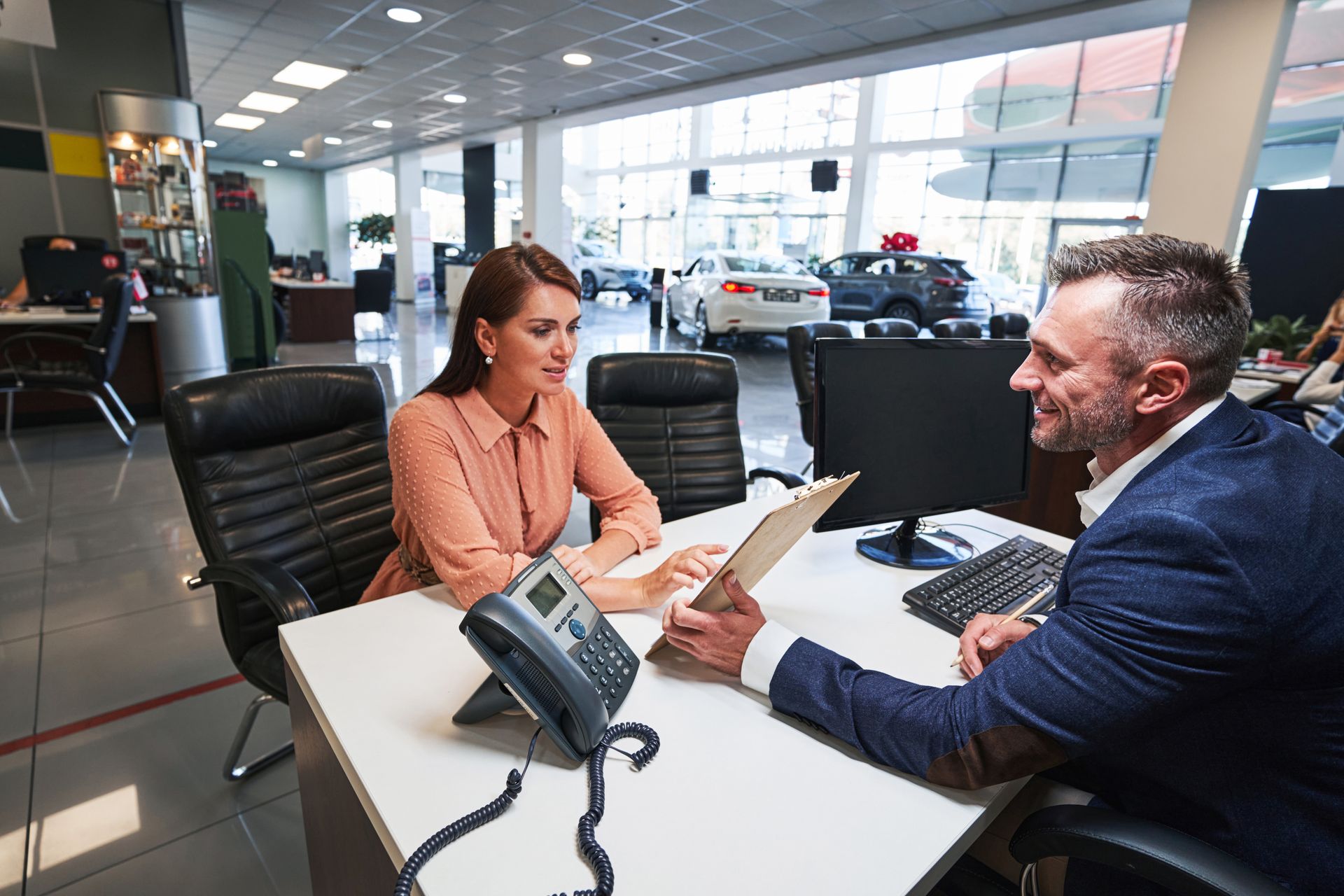 A man and a woman are sitting at a desk in a car dealership.