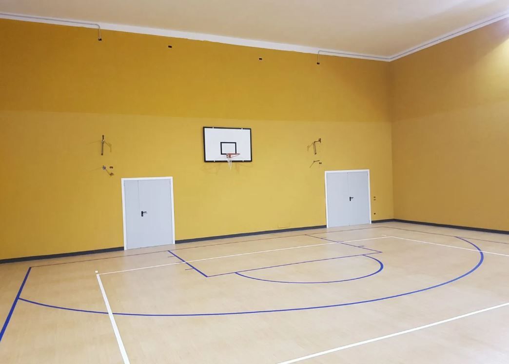 Campo da basket in una palestra con pareti gialle, canestro e due porte chiuse.