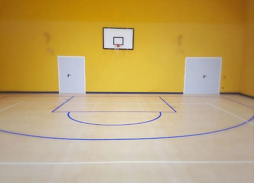 Campo da basket con muro giallo, canestro, linee blu, due porte bianche.