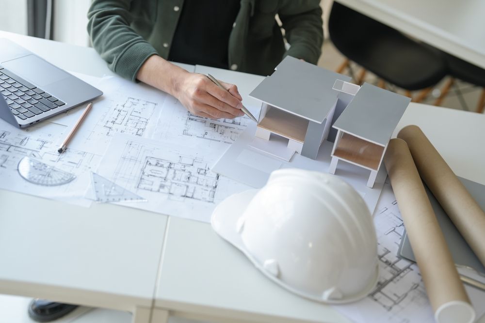 Architect working on blueprints at a desk, with a model house, hard hat, and rolled plans.